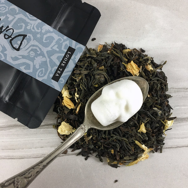 Black bag with grey spooky vine graphics, DemBones Jasmine Tea in pile with skull sugar cubes on old silver spoon